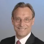 Head-and-shoulders portrait of a smiling man in a dark suit and red tie, wearing glasses against a blue gradient background.