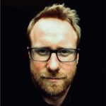 Portrait of a man with short blond hair and a light beard wearing black rectangular glasses against a dark background, looking at the camera.